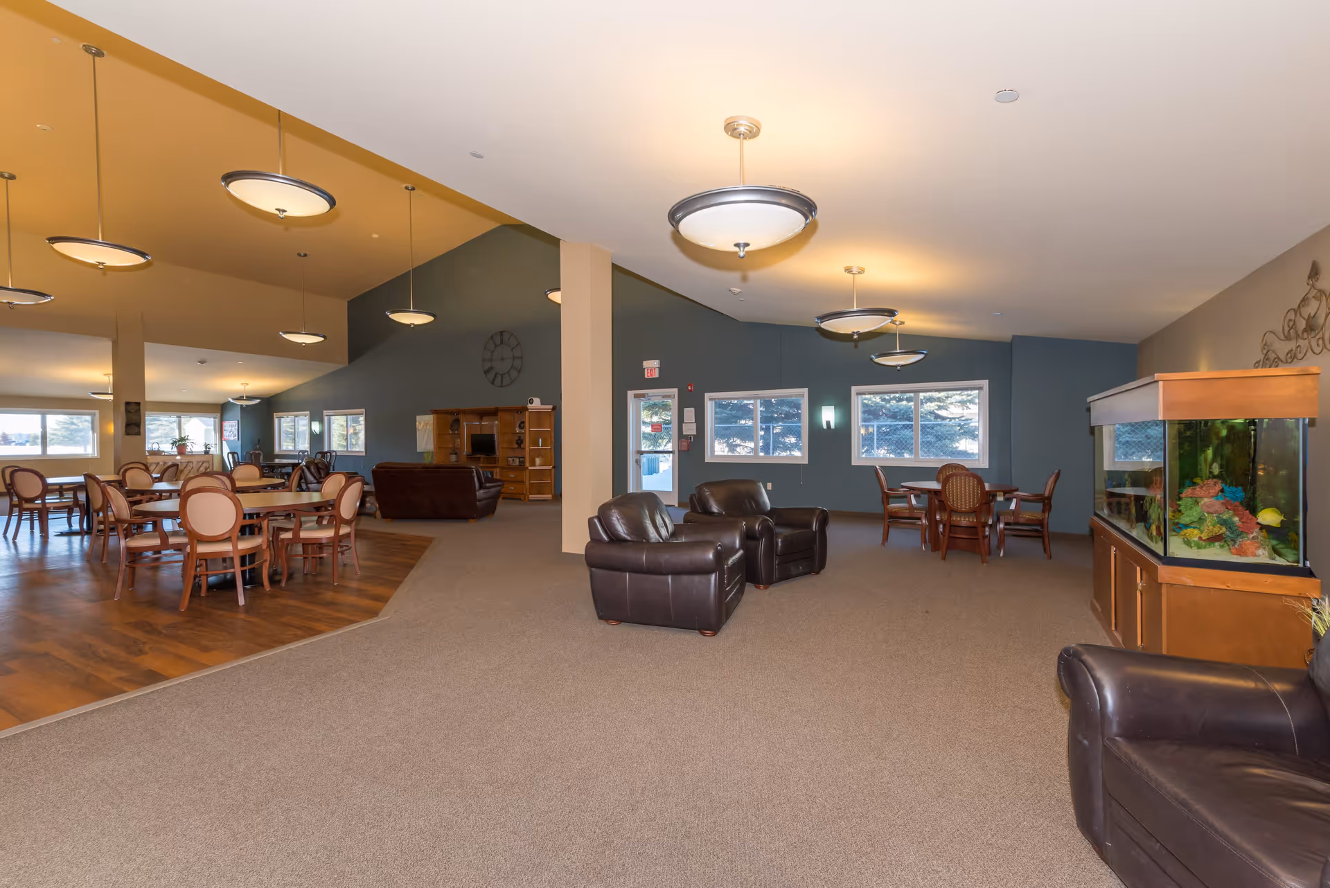 Spacious common area in a senior living facility featuring multiple seating arrangements including round dining tables with chairs, leather armchairs, and a large fish tank. The room has a mix of carpet and wood flooring, large windows letting in natural light, and several ceiling lights.