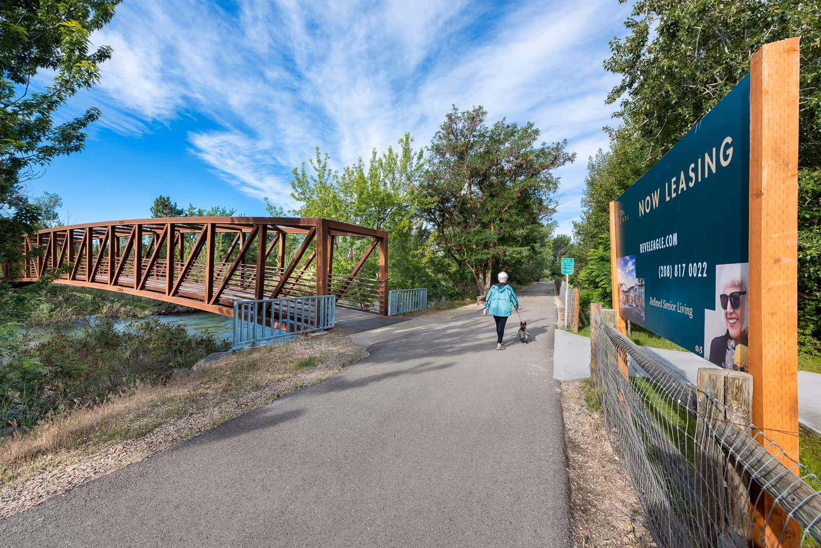 A person walking a small dog on a paved path next to a wooden and metal pedestrian bridge over a river. There are trees and greenery on both sides of the path, and a large sign on the right side that reads 'NOW LEASING' with contact information for Revel Eagle, a senior living facility.