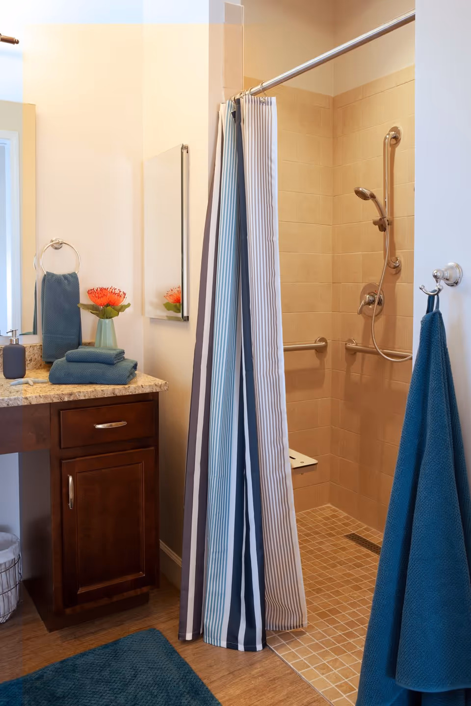 A bathroom with a walk-in shower featuring beige tiles, a handheld showerhead, and grab bars. The shower has a striped curtain partially open. Next to the shower is a wooden vanity with a granite countertop, a mirror, a blue towel hanging on a ring, folded blue towels on the counter, a soap dispenser, and a vase with an orange flower. A blue bath mat is on the wooden floor, and a blue towel hangs on a hook on the wall.