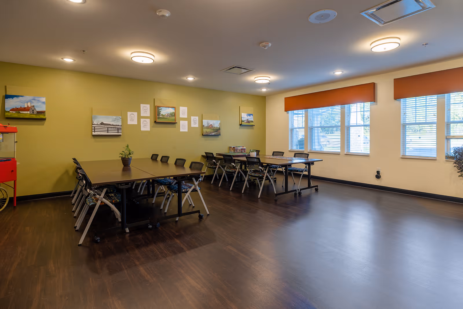 A spacious room with two long tables surrounded by chairs, a green accent wall decorated with paintings and sketches, large windows with orange valances letting in natural light, and a red popcorn machine in the corner.