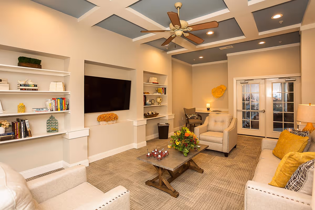 A cozy living room with beige walls and carpet, featuring a large flat-screen TV mounted on the wall between built-in shelves filled with books and decorative items. The room has comfortable beige armchairs and a sofa with yellow and patterned cushions, a wooden coffee table with a floral arrangement and small desserts, a ceiling fan with wooden blades, and double glass doors at the back.