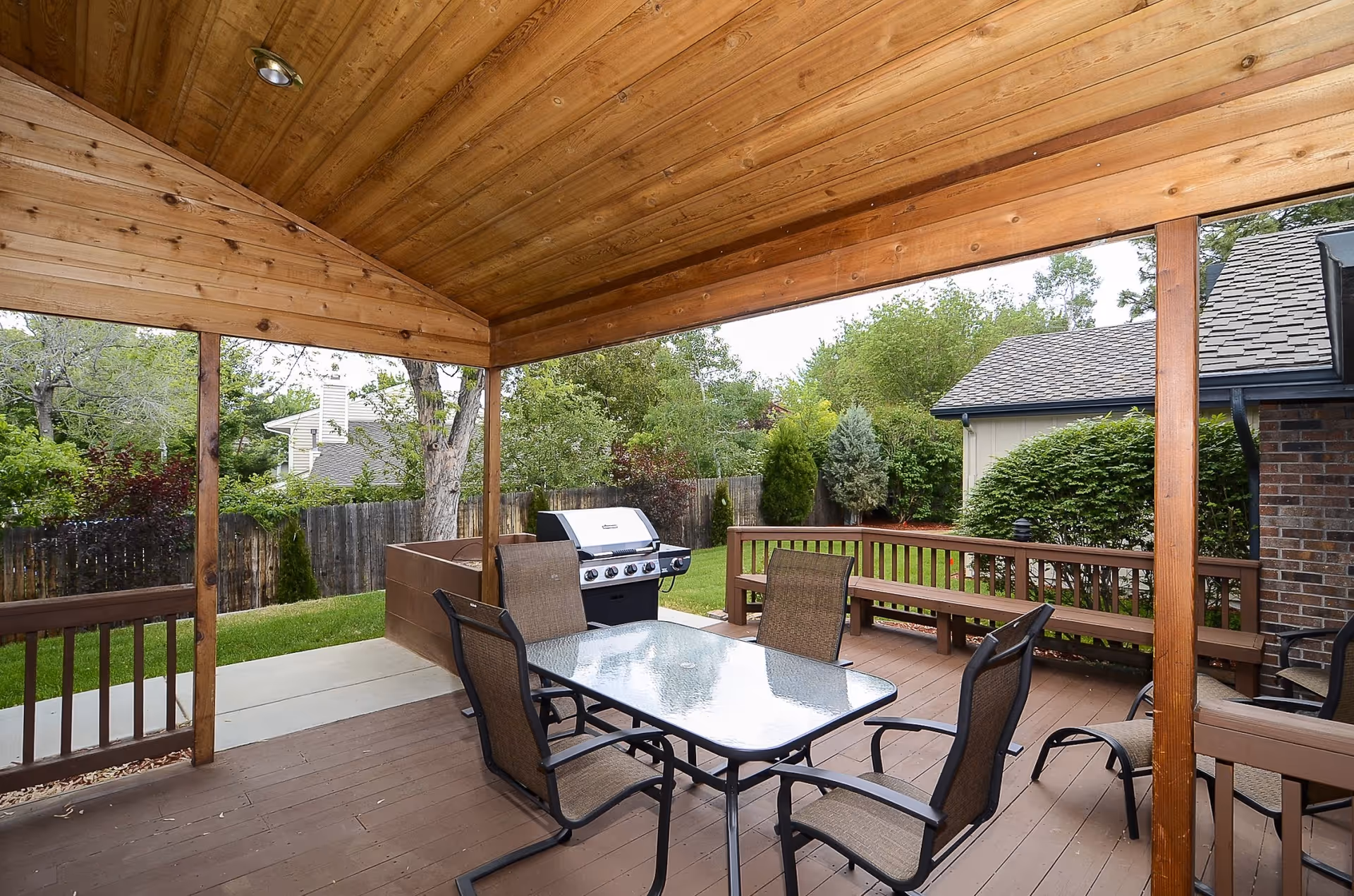 Covered outdoor patio area with a wooden ceiling and floor, featuring a glass-top table with four chairs, a built-in bench along the railing, and a gas grill. The patio overlooks a fenced backyard with green grass, trees, and shrubs.