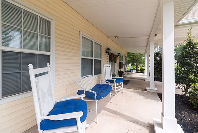 A covered outdoor porch area with white rocking chairs and a white metal bench with blue cushions. The porch has beige siding, white pillars, and windows with blinds. There are plants and greenery visible at the edge of the porch.