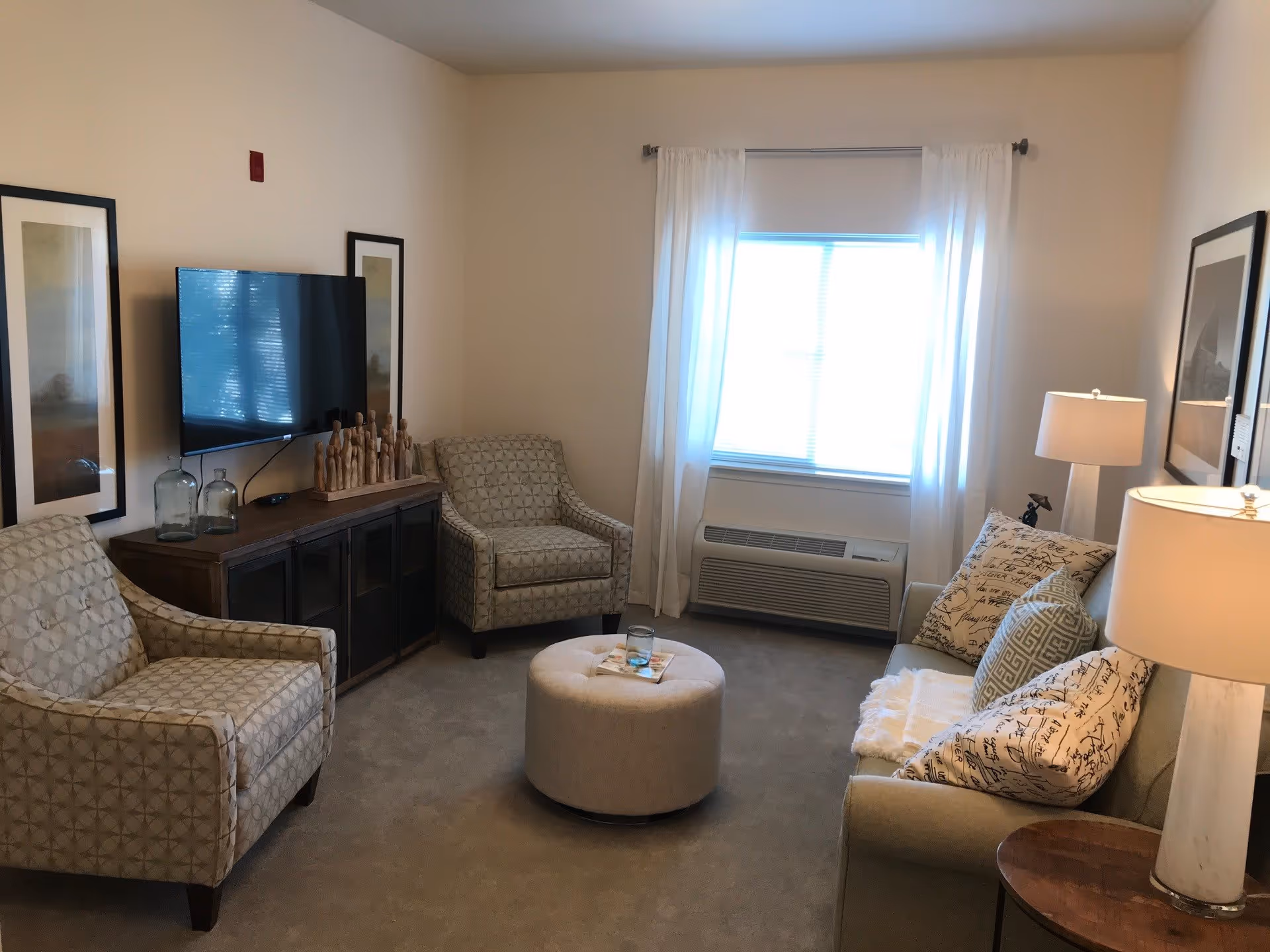 A cozy living room with two patterned armchairs, a beige sofa with decorative pillows, a round ottoman in the center, a wooden TV stand with a flat-screen television mounted above it, two framed pictures on the wall, a window with white curtains, and two table lamps providing warm lighting.