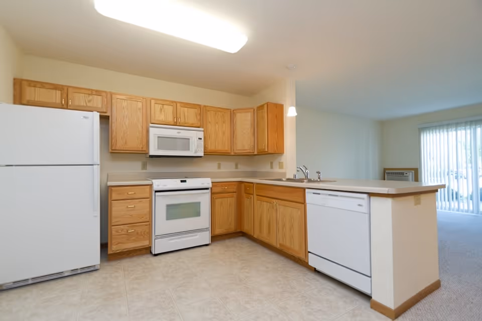 A clean and bright kitchen with wooden cabinets, a white refrigerator, white stove with oven, white microwave above the stove, and a white dishwasher. The kitchen has a beige countertop with a double sink and a hanging light fixture above it. The floor is tiled, and the kitchen opens into a carpeted living area with vertical blinds covering a sliding glass door.