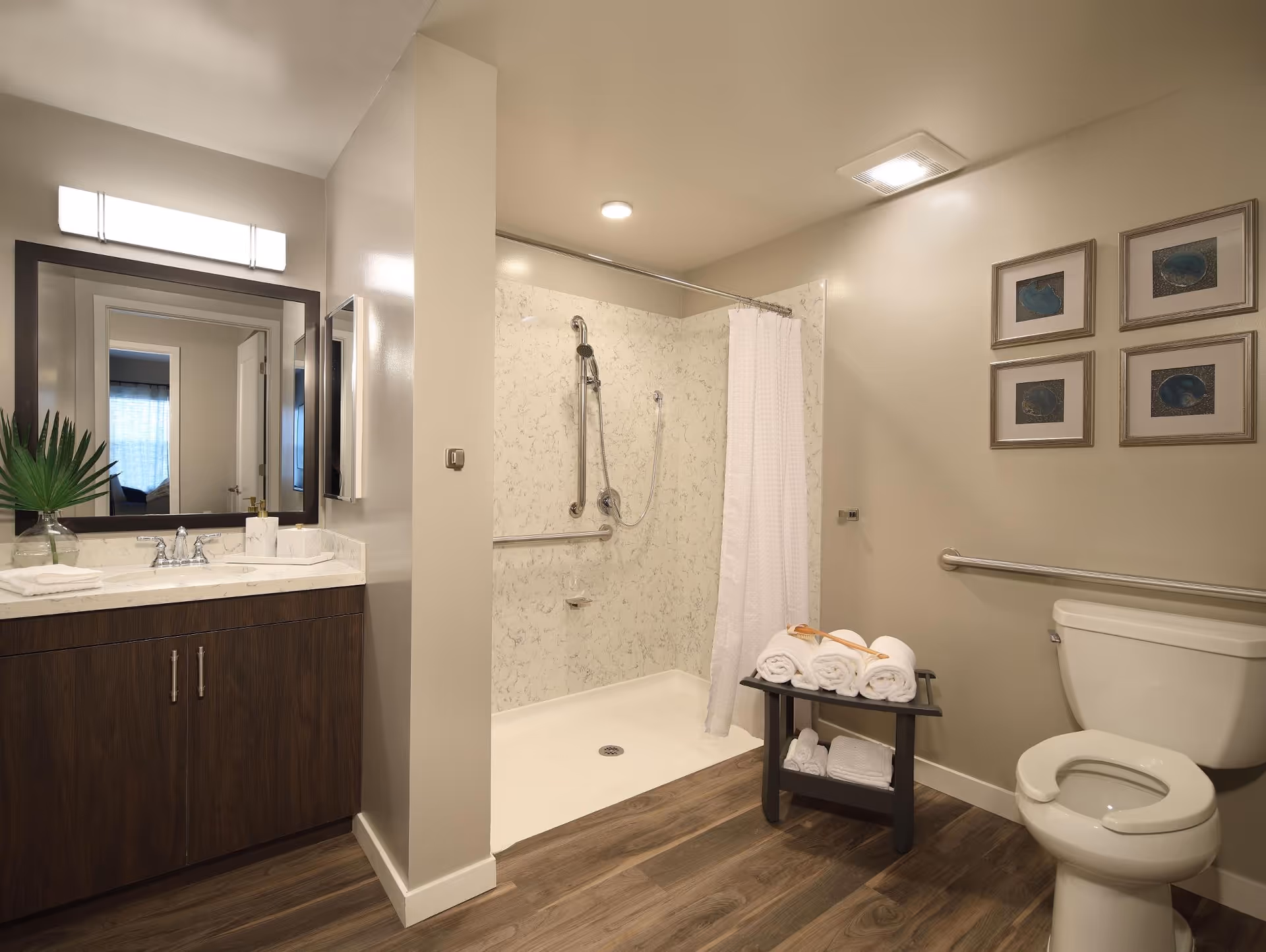 Accessible bathroom featuring a walk-in shower with grab bars, vanity with mirror, toilet and a small table of rolled towels.