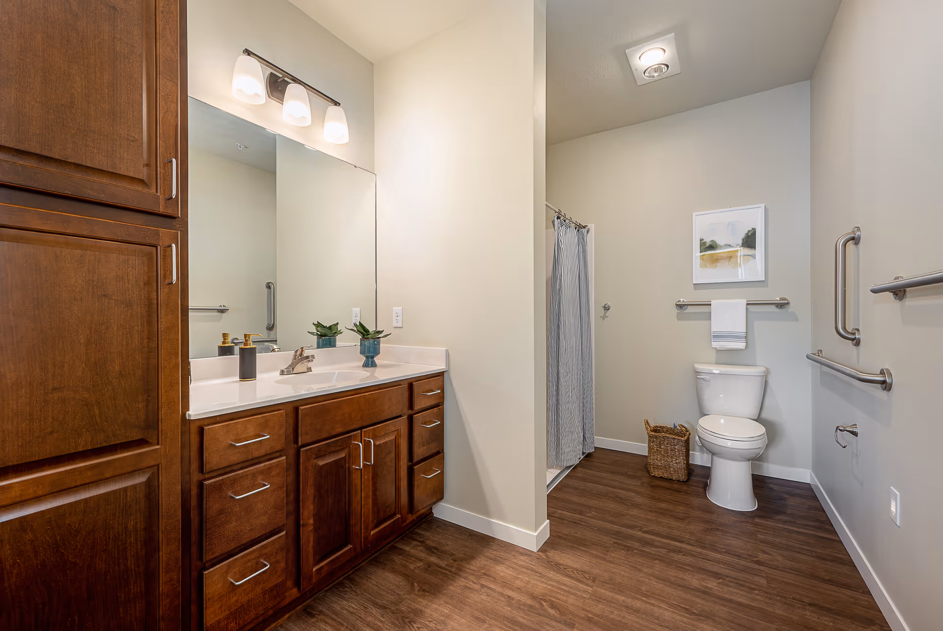 A clean and modern bathroom featuring a large mirror above a wooden vanity with multiple drawers and cabinets. The vanity has a white countertop with a sink, soap dispenser, and a small plant. To the right, there is a toilet with a towel hanging above it, a wicker basket on the floor, and a shower area with a curtain. The bathroom has light-colored walls, wood-style flooring, and several grab bars for accessibility.