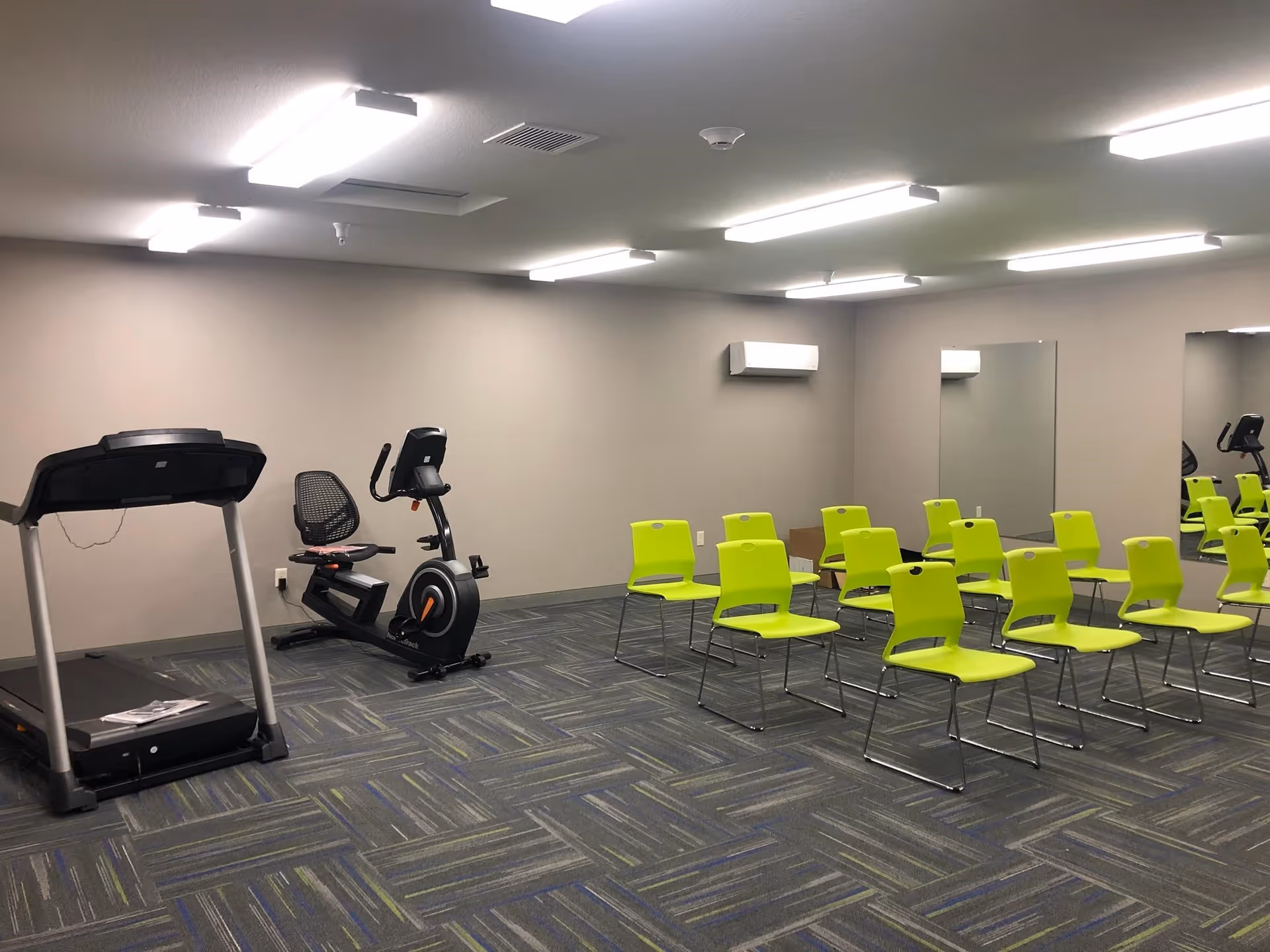 A room with exercise equipment including a treadmill and a stationary bike on the left side, and several lime green chairs arranged in rows on the right side. The room has gray walls, a patterned carpet floor, bright overhead fluorescent lighting, and two large mirrors on the right wall.