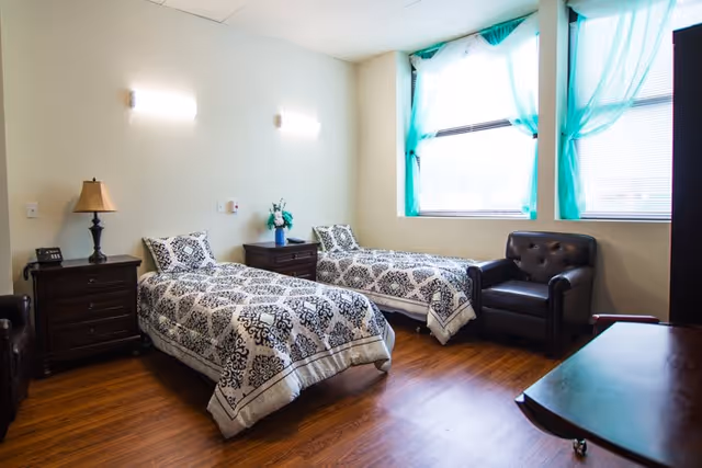 A well-lit bedroom with two single beds featuring patterned bedspreads, a wooden nightstand with a lamp and a flower vase between the beds, a dark leather armchair near two large windows with turquoise curtains, and wooden flooring.