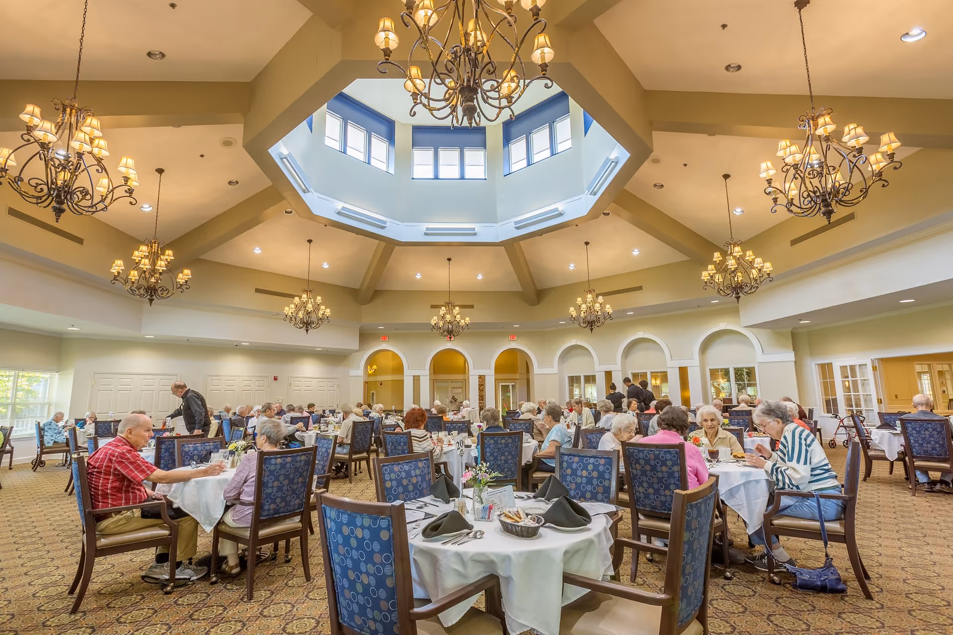 A spacious dining room with a high ceiling and multiple chandeliers. Several elderly people are seated at round tables covered with white tablecloths, engaged in conversation and dining. The room has large windows and an octagonal skylight that lets in natural light.