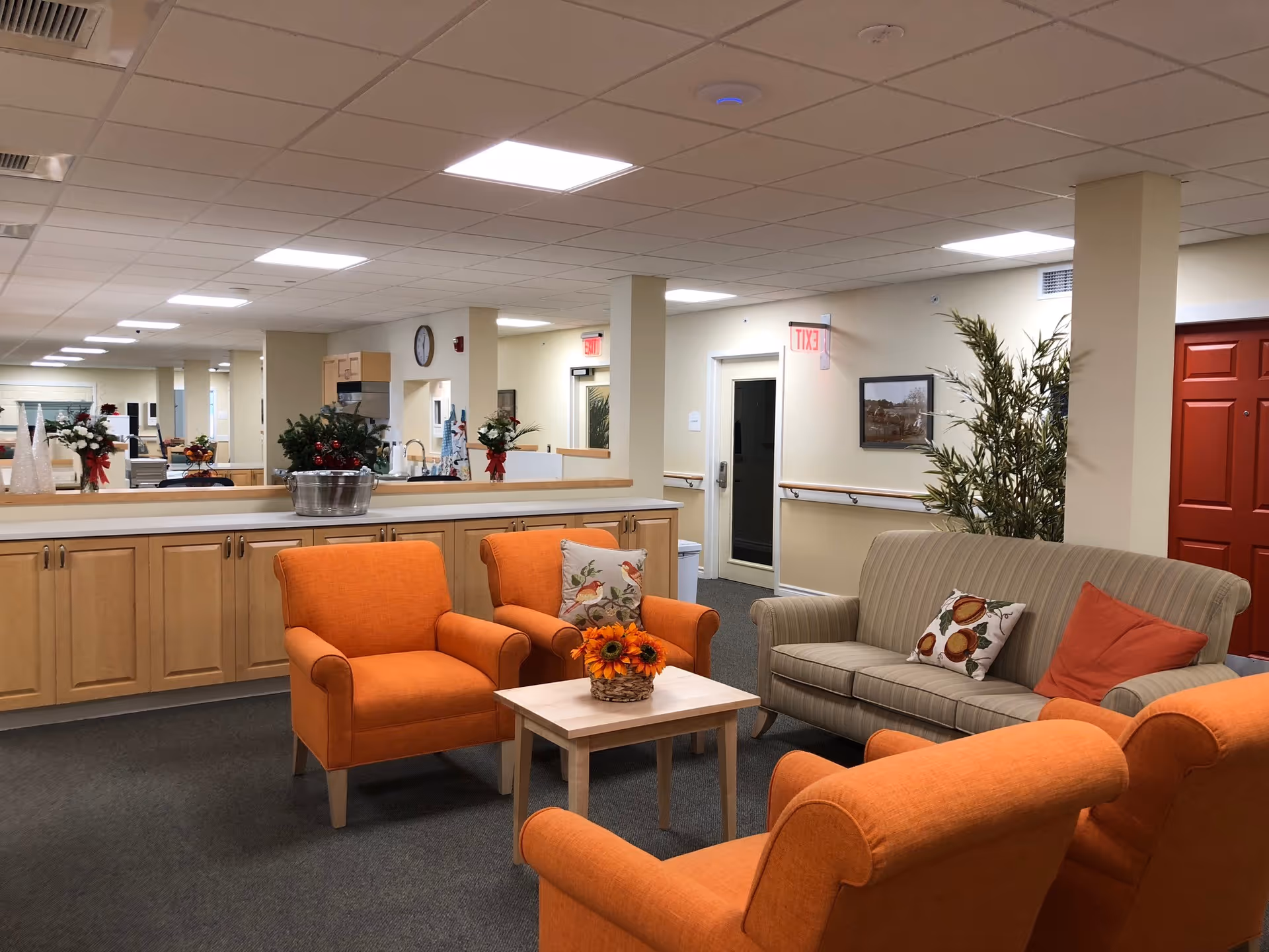A cozy common area in a senior living facility with three orange armchairs and a beige striped sofa arranged around a small wooden coffee table with a basket of sunflowers. The room has light-colored walls, a red door, potted plants, and a long counter with cabinets underneath. The ceiling has recessed lighting, and there is a clock and framed artwork on the walls.