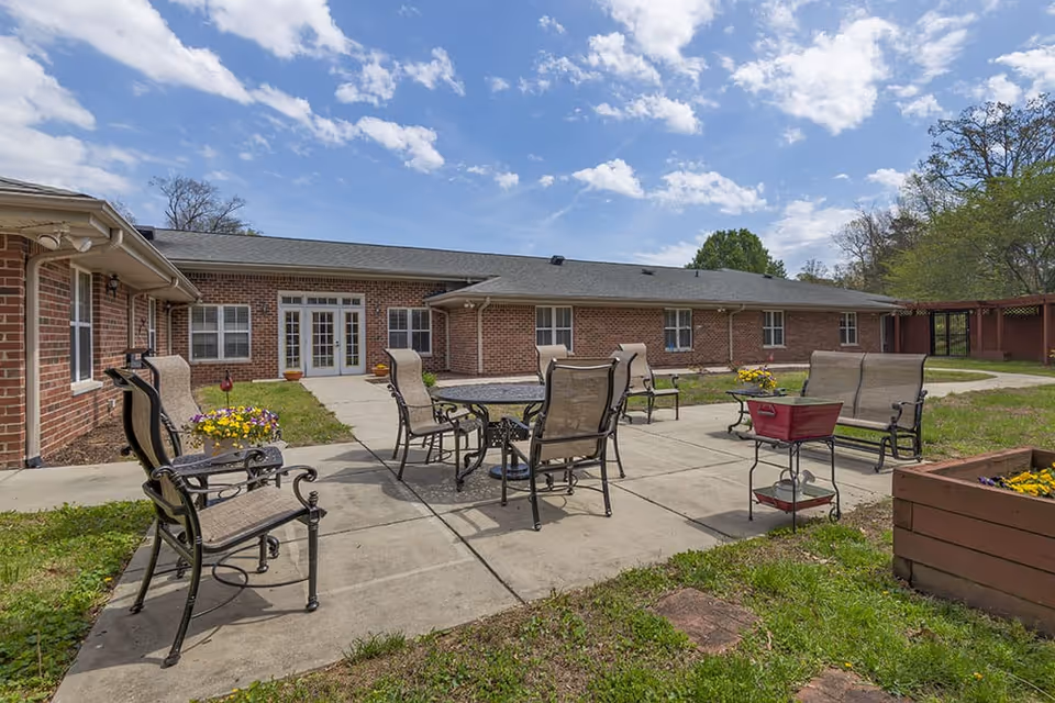 Outdoor patio area at Brookdale High Point North featuring metal chairs and tables arranged on a concrete surface, surrounded by grass and flower planters, with a brick building and blue sky with clouds in the background.