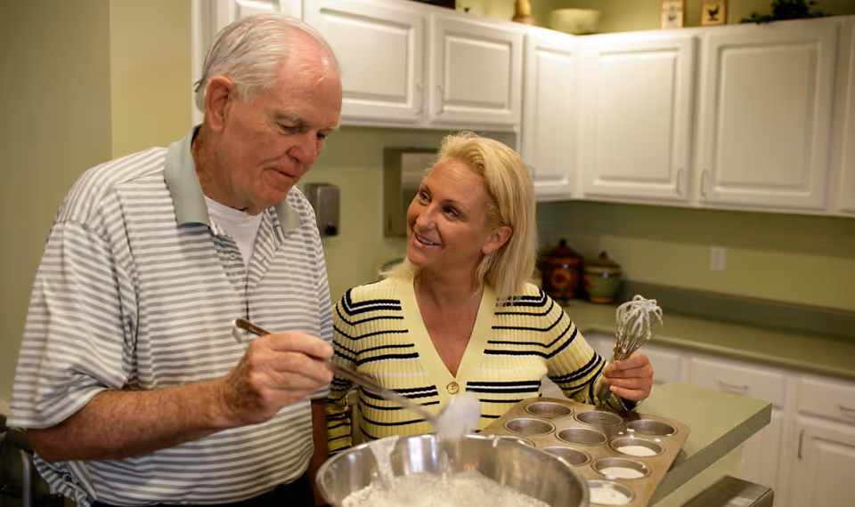 An elderly man and a middle-aged woman are in a kitchen preparing food together. The man is scooping batter from a large mixing bowl into a muffin tin, while the woman holds a whisk and looks at him with a smile. The kitchen has white cabinets and a green countertop.