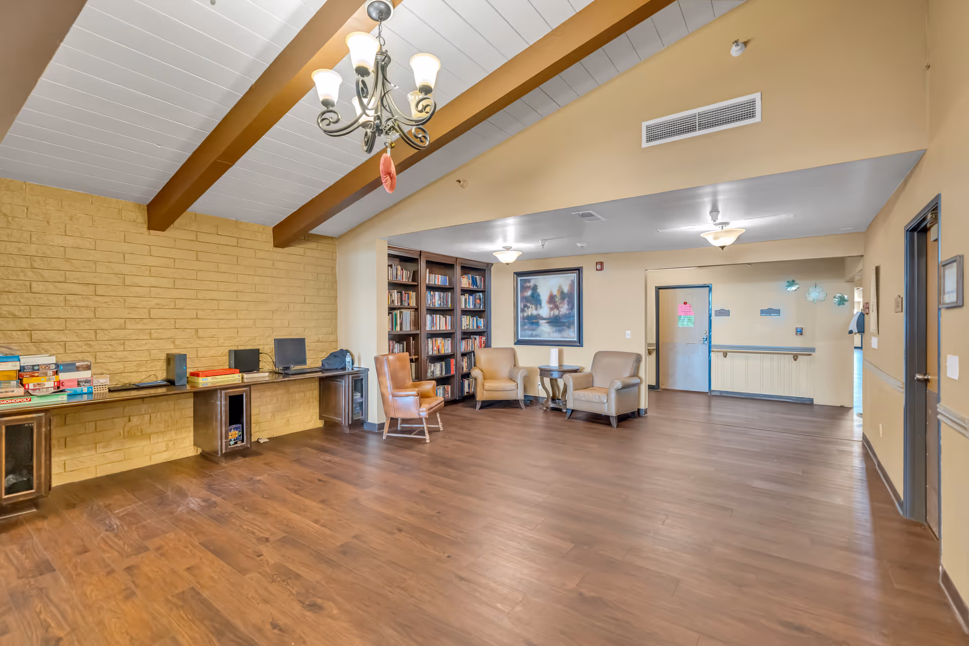 A spacious common area with wooden flooring and a high ceiling with exposed beams and a chandelier. There is a built-in bookshelf filled with books, a few armchairs arranged around a small table, and a long desk against a yellow brick wall with board games and a computer. The room has beige walls and doors leading to other areas.