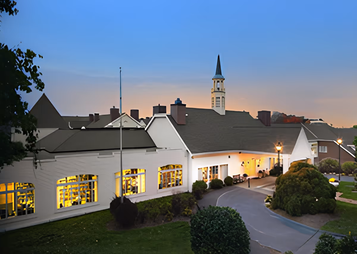 Exterior view of a senior living facility building at dusk with illuminated windows, a tall steeple, and a curved driveway surrounded by landscaped bushes and trees.