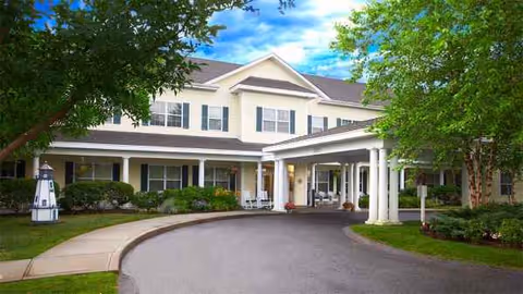 Two-story senior living facility entrance with circular driveway, columns, and surrounding trees.