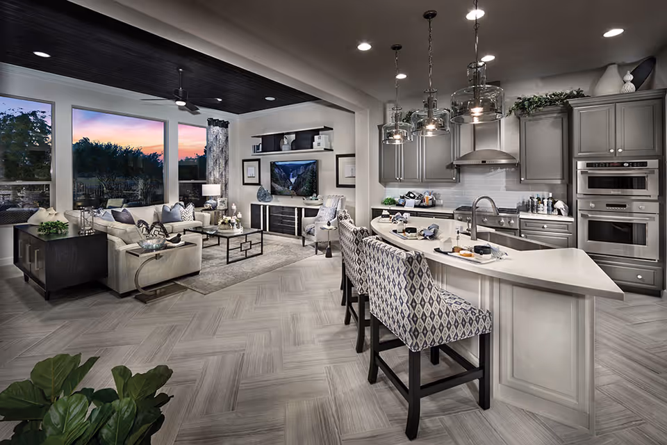 Modern open-concept living room and kitchen area with large windows showing a sunset outside. The living room has a beige sectional sofa, a coffee table, a TV mounted on a wall with shelves, and decorative items. The kitchen features gray cabinetry, a white island with a sink, three patterned bar stools, and pendant lights hanging above the island.