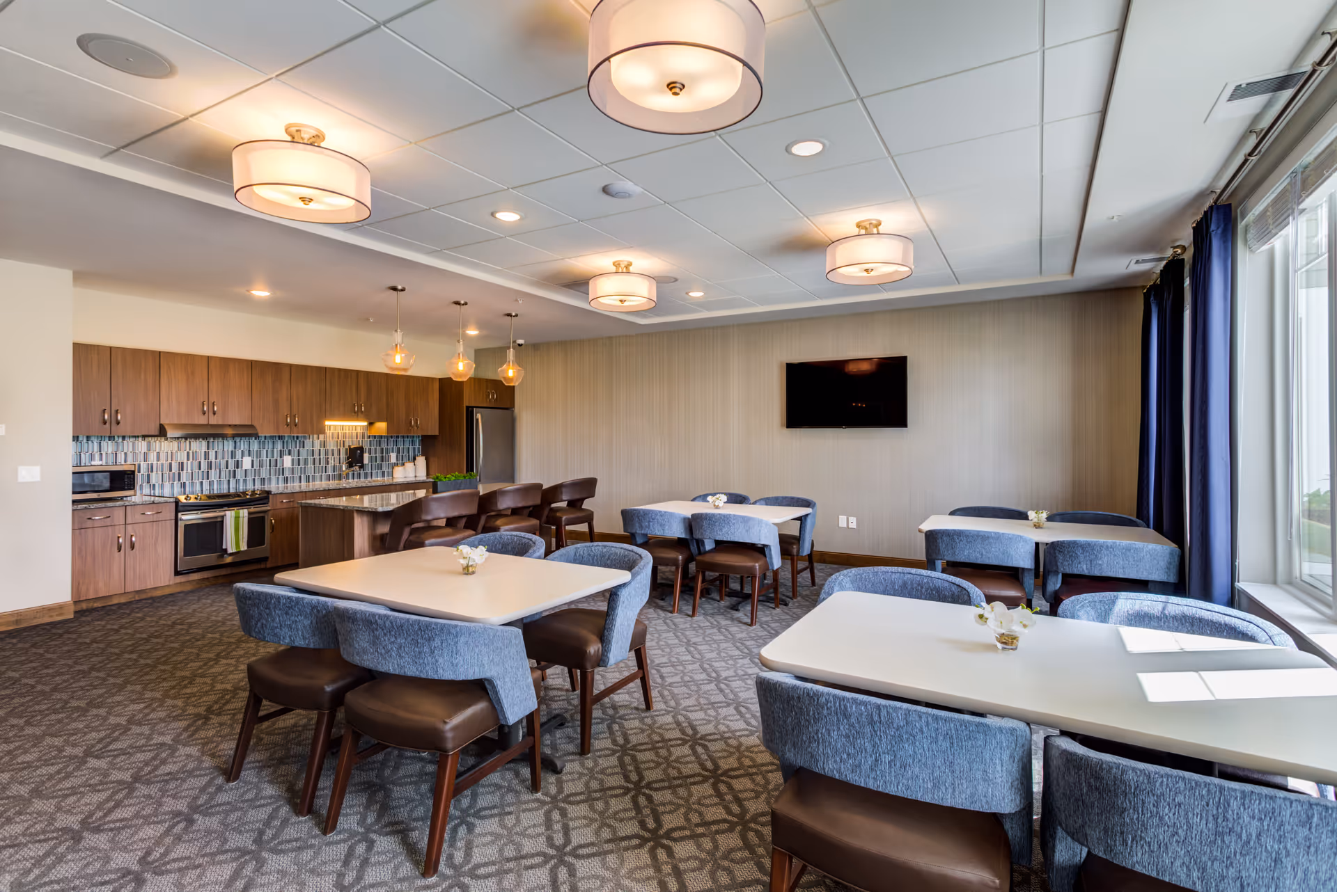 A bright and modern dining area in a senior living facility featuring several tables with blue upholstered chairs and small flower arrangements. The room includes a kitchen area with wooden cabinets, a stainless steel refrigerator, stove, microwave, and a kitchen island with bar stools. Large windows with dark curtains allow natural light to fill the space, and a flat-screen TV is mounted on the wall.