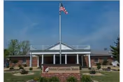 Front entrance of Aviston Countryside Manor brick building with an American flag on a flagpole and landscaped grounds.