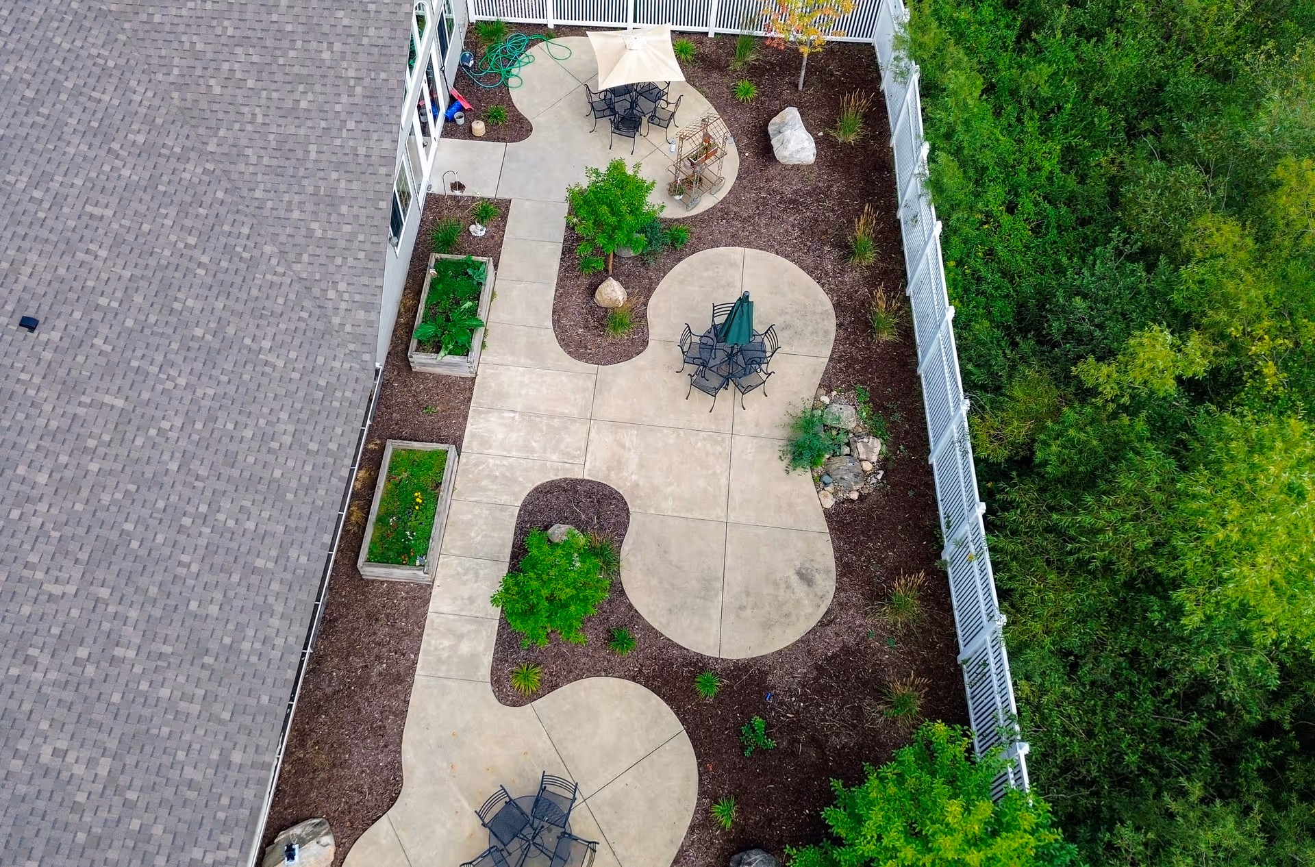 Aerial view of a landscaped outdoor patio area at Golden Horizons of Aitkin, featuring curved concrete walkways, several seating areas with tables and chairs, garden beds with plants, small trees, and a white fence bordering the space.
