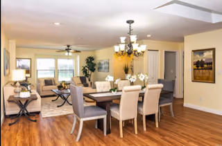 Open-plan dining and living area with a wooden dining table and upholstered chairs in front of a sitting area with sofas and large windows.