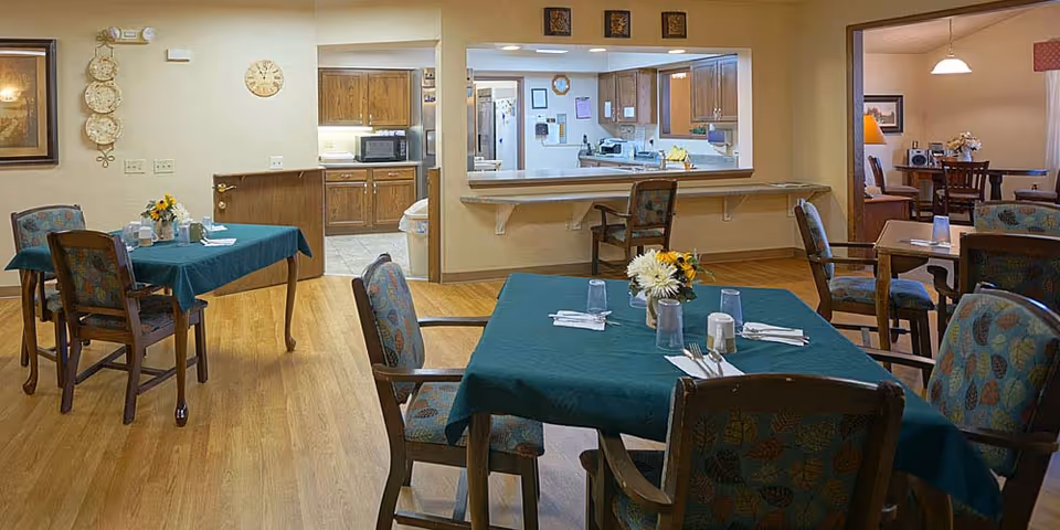 A dining area in a senior living facility with several tables covered in green tablecloths, each set with glasses, napkins, and utensils. The room has wooden floors and patterned chairs. In the background, there is a kitchen area visible through a large pass-through window, and another dining space is partially visible through an open doorway.