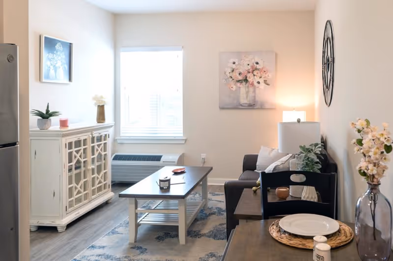 A cozy living room with a dark gray sofa adorned with pillows, a wooden coffee table with a candle and small decor items, a white cabinet with glass doors, a window with blinds, a wall clock, and floral artwork on the walls. A dining table with a plate, placemat, and flowers in a vase is partially visible in the foreground.