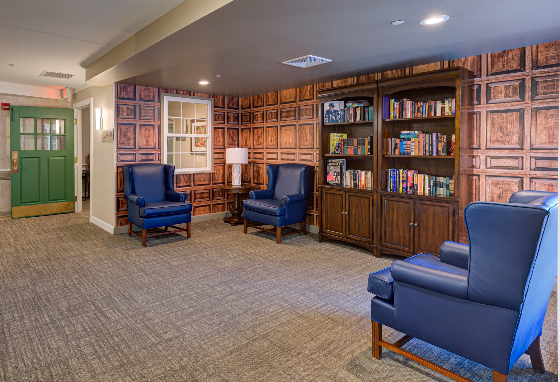 A cozy interior common area with three blue leather armchairs arranged around a wooden side table with a lamp. The walls have a decorative wood panel pattern, and there is a large wooden bookshelf filled with books. A green door and a window are visible in the background.