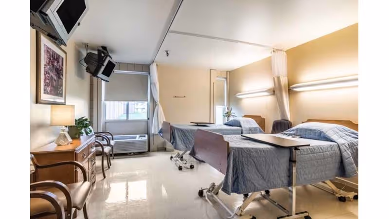 A clean and well-lit hospital or care facility room with two adjustable beds covered in blue quilts. Each bed has an overbed table. There are two chairs and a wooden dresser with a lamp and a plant on it. A window with a roller shade and an air conditioning unit is visible. Two televisions are mounted on the wall near the ceiling.
