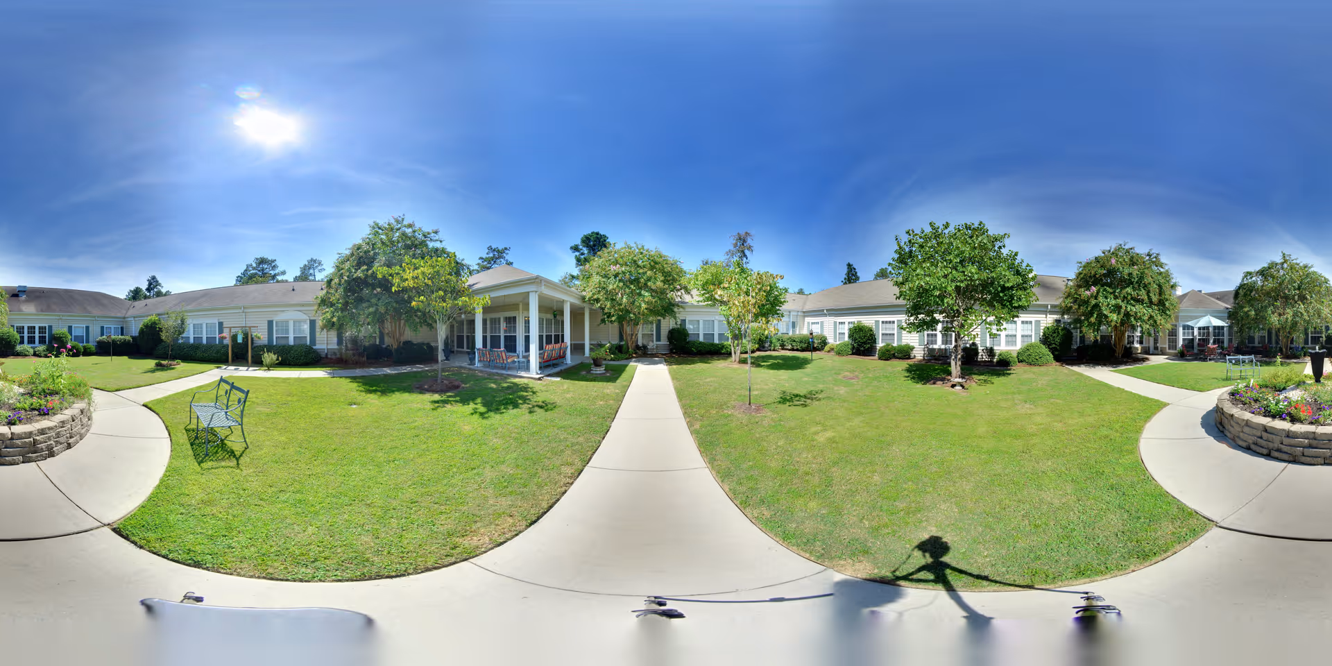 Wide panoramic view of a senior living facility courtyard with a well-maintained lawn, several trees, flower beds bordered by stone walls, a few benches, and a covered porch area attached to a single-story building under a clear blue sky.