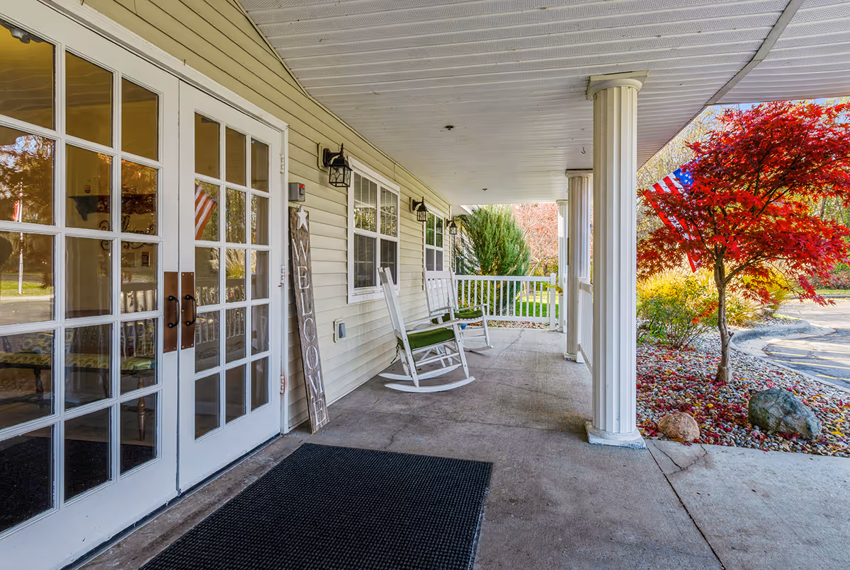 Covered porch area with white rocking chairs, a welcome sign leaning against the wall, white double doors with glass panes, and a view of a red-leaved tree and American flag outside.