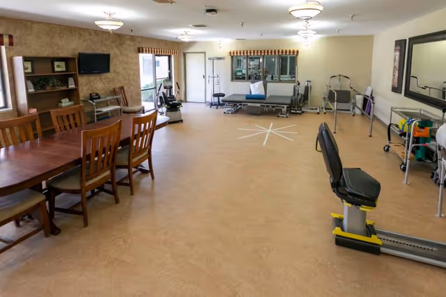 A spacious rehabilitation room with exercise equipment including a recumbent bike, parallel bars, and therapy tables. There is a wooden dining table with chairs on the left side, a TV mounted on the wall, and large windows with striped valances letting in natural light. The room has beige flooring and light-colored walls with ceiling lights.