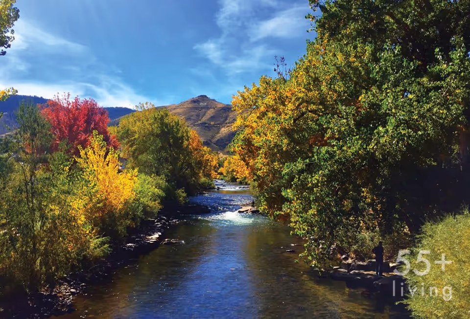 A scenic outdoor view of a river flowing through a landscape with trees showing autumn colors of red, yellow, and green, with hills in the background under a partly cloudy blue sky.