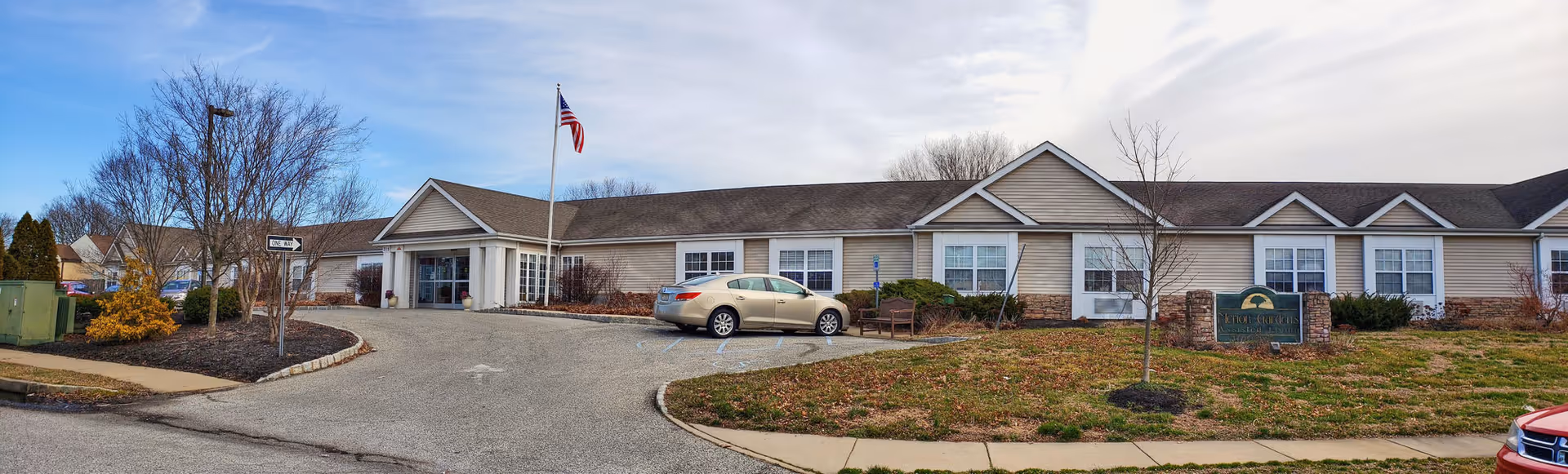 Front exterior view of Merion Gardens Assisted Living facility with a single-story building, an American flag on a flagpole, a parked car, and a sign with the facility name on a grassy area.