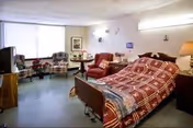 A senior living facility room with a hospital-style bed covered in a red and white patterned blanket. The room includes a red armchair, a small round table with a lamp, two upholstered chairs near a large window with sheer curtains, a television on a wooden stand, and framed artwork on the walls.