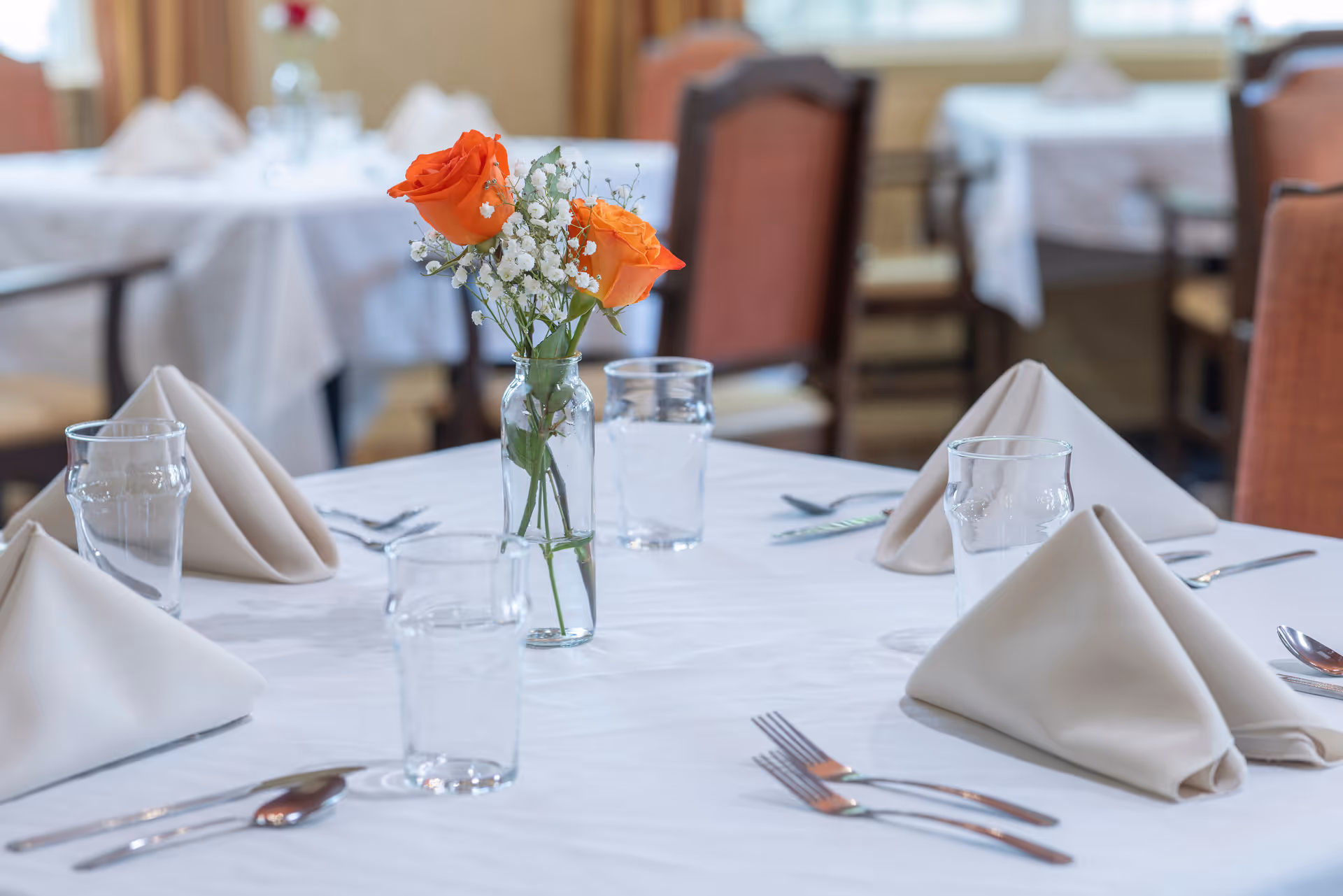 A dining table set with white tablecloth, beige folded napkins, clear drinking glasses, silverware, and a small glass vase holding two orange roses and white baby's breath flowers. In the background, other similarly set tables and chairs are visible in a softly lit dining room.