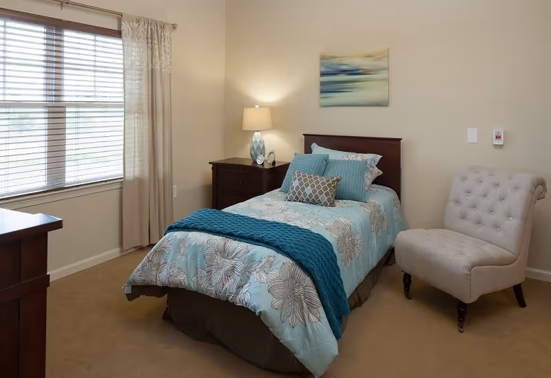 A tidy single bedroom with a floral-patterned bed dressed in teal accents, a nightstand with lamp, an upholstered chair, and a window with blinds.