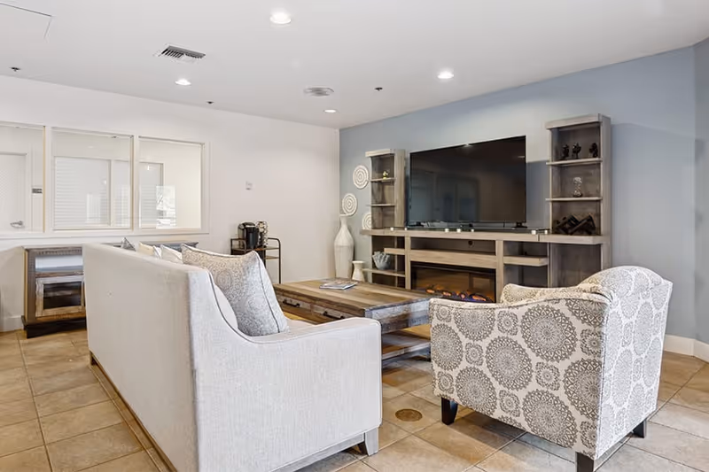 A cozy living room area with a light gray sofa and a patterned armchair facing a large flat-screen TV mounted on a wooden entertainment center. The room has tiled flooring, a wooden coffee table, decorative vases, and shelves with small decor items. The walls are painted light blue and white, and there are recessed ceiling lights.
