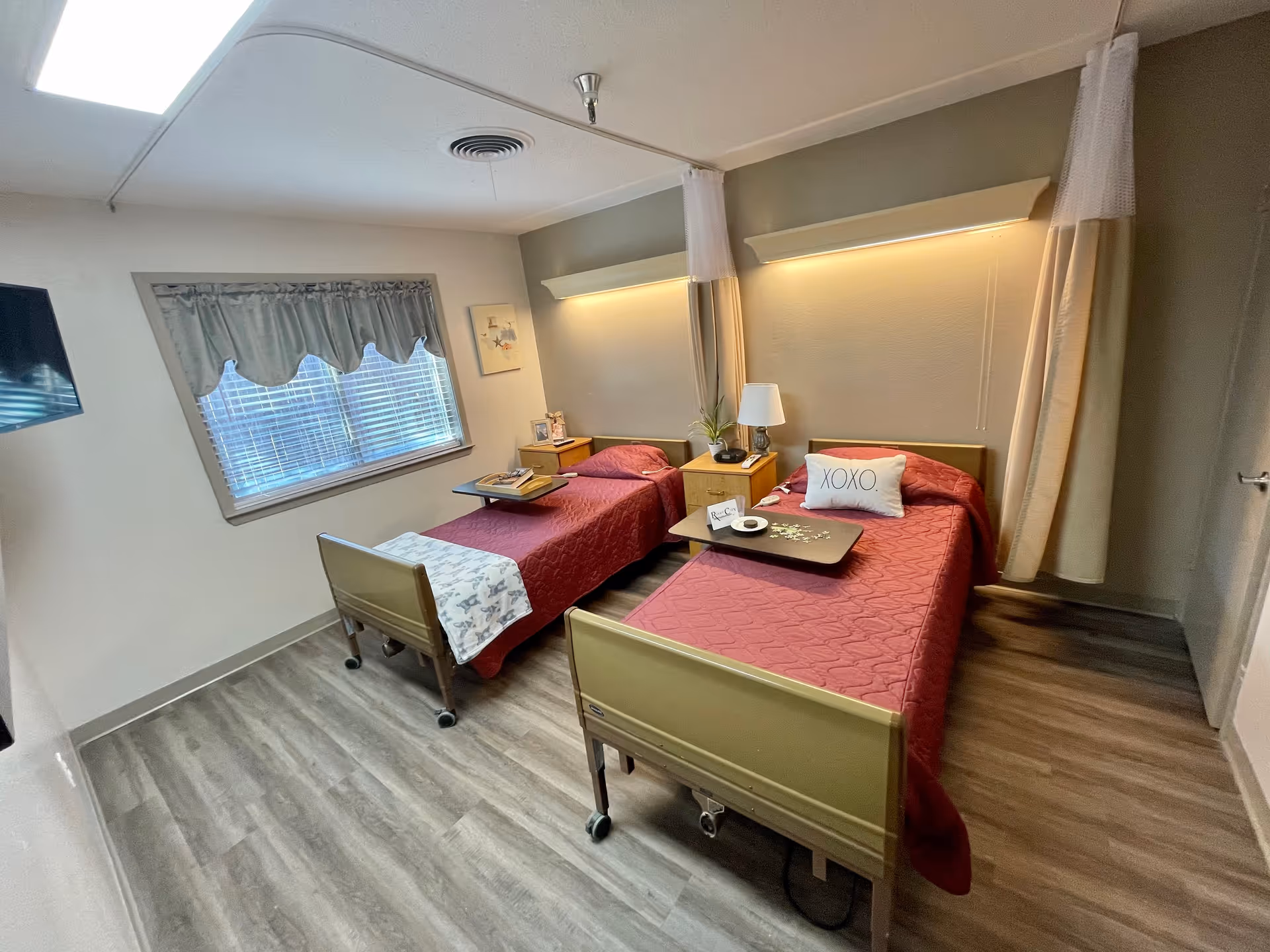 Shared bedroom with two hospital-style twin beds with red bedspreads, a bedside table and lamp, window with valance, and wood-look flooring.