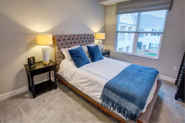 A neatly made bed with a tufted headboard in a bedroom. The bed has white sheets, two blue pillows, and a blue throw blanket. There are two black nightstands on either side of the bed, each with a lamp and a small decorative item. A large window with a view of neighboring buildings is on the right side of the room.