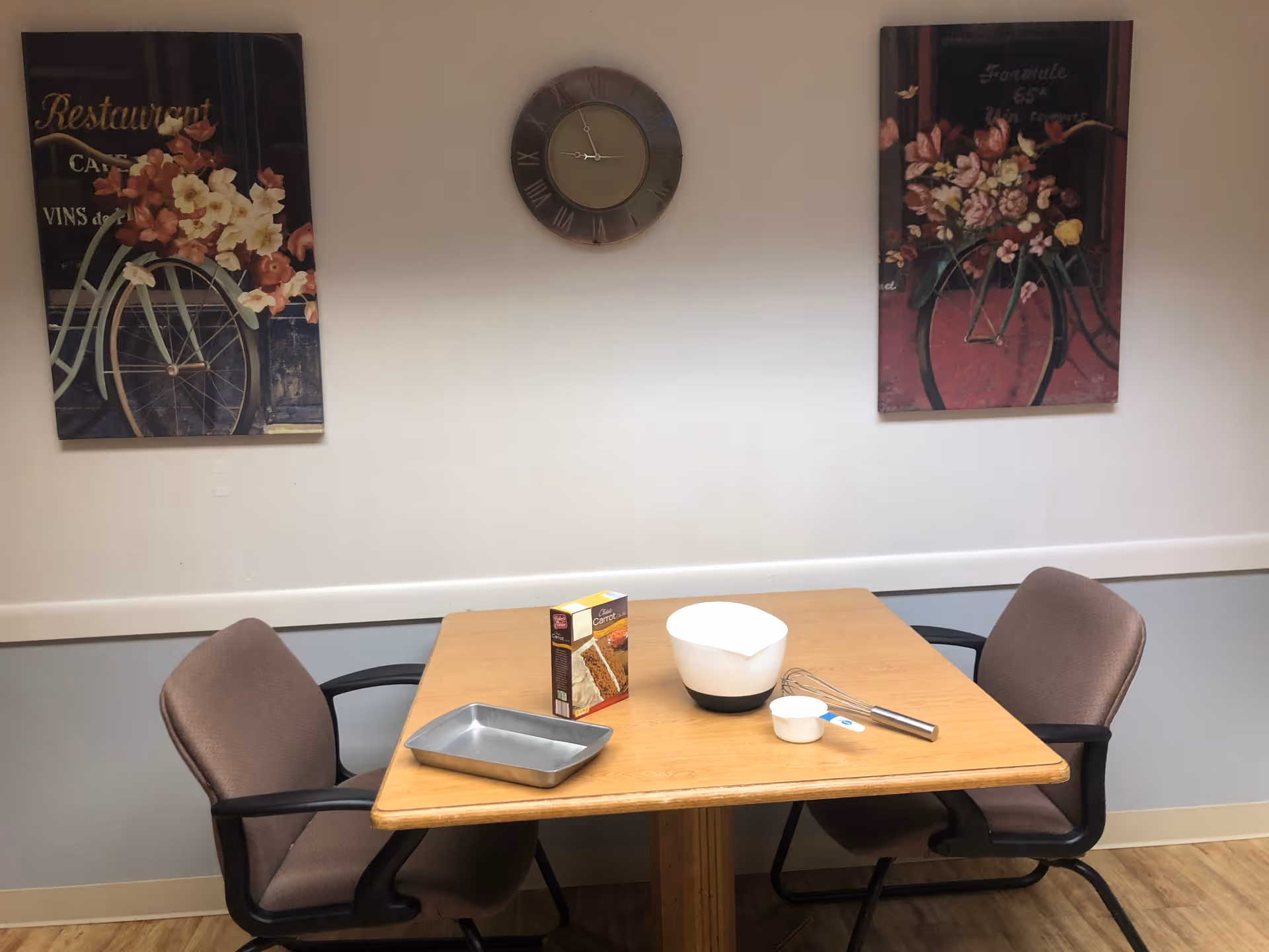 A small dining area with a wooden table and two brown cushioned chairs. On the table are a baking pan, a box of carrot cake mix, a white mixing bowl, a metal whisk, and a measuring cup. The wall behind the table has a clock in the center and two paintings featuring bicycles with flowers on either side.