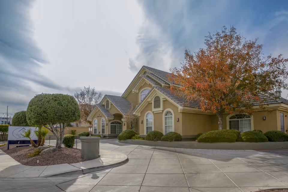 Exterior view of Villa Court Assisted Living facility showing a beige building with multiple peaked roofs, arched windows, and well-maintained landscaping including trimmed bushes and a tree with autumn-colored leaves under a partly cloudy sky.