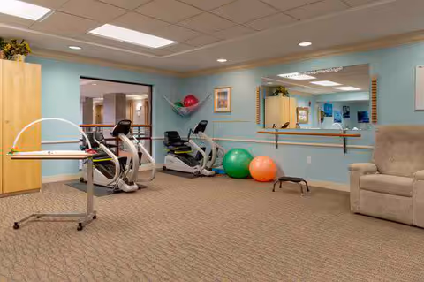 A small fitness/therapy room with two recumbent exercise bikes, colorful therapy balls, a wall mirror with a barre, and a recliner.