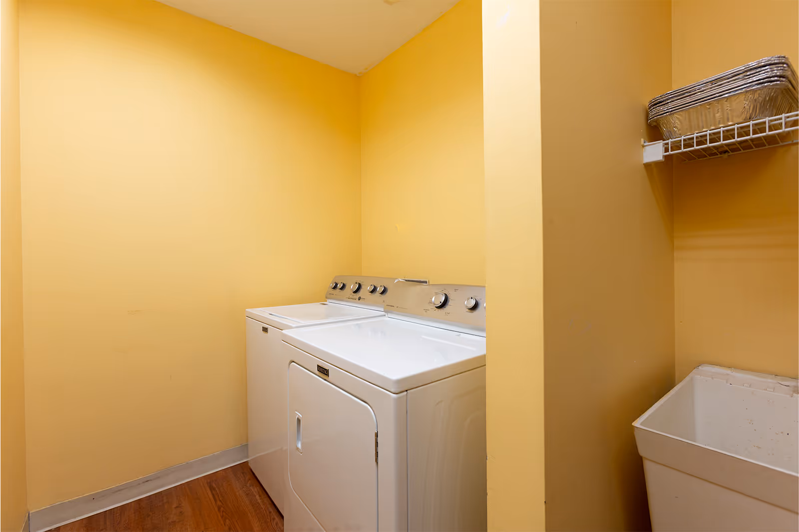 Small laundry room with a washer and dryer against yellow walls, a utility sink and a wire shelf with a basket.