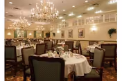 A spacious dining room with multiple round tables covered with white tablecloths, set with plates, glasses, and silverware. The room features elegant chandeliers hanging from the ceiling, framed artwork on the walls, and upholstered chairs around each table. The overall ambiance is warm and inviting.