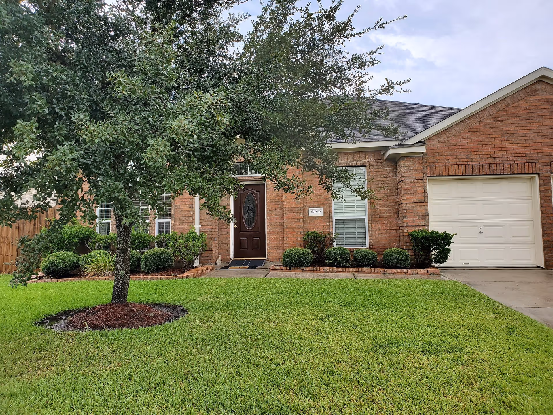 Front exterior view of a single-story brick building with a dark brown front door, white garage door, several windows with white blinds, neatly trimmed bushes, a tree in the front yard, and a well-maintained green lawn.