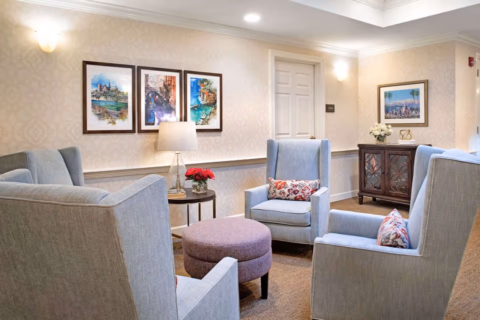A cozy sitting area in a senior living facility with four light blue upholstered armchairs arranged around a round purple ottoman. A small side table with a lamp and a vase of red flowers is next to one chair. The walls are decorated with framed artwork and a wooden cabinet with decorative items is visible in the background.