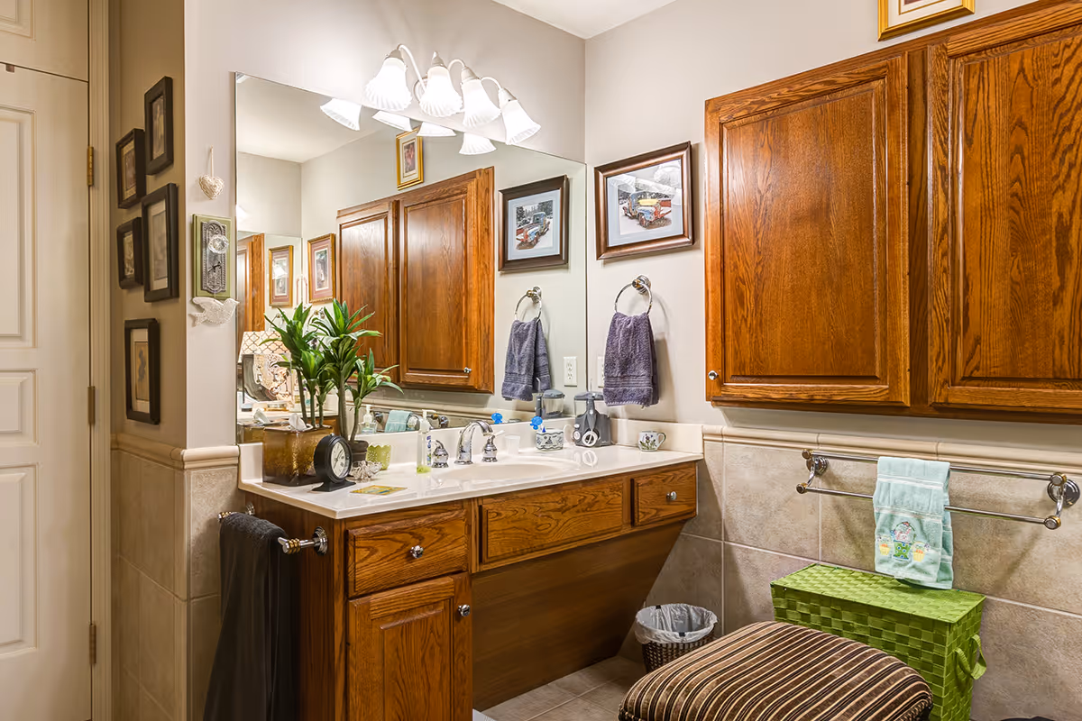 Well-lit bathroom vanity with wooden cabinets, a large mirror, sink, towels, and decorative items including a green storage basket and striped stool.