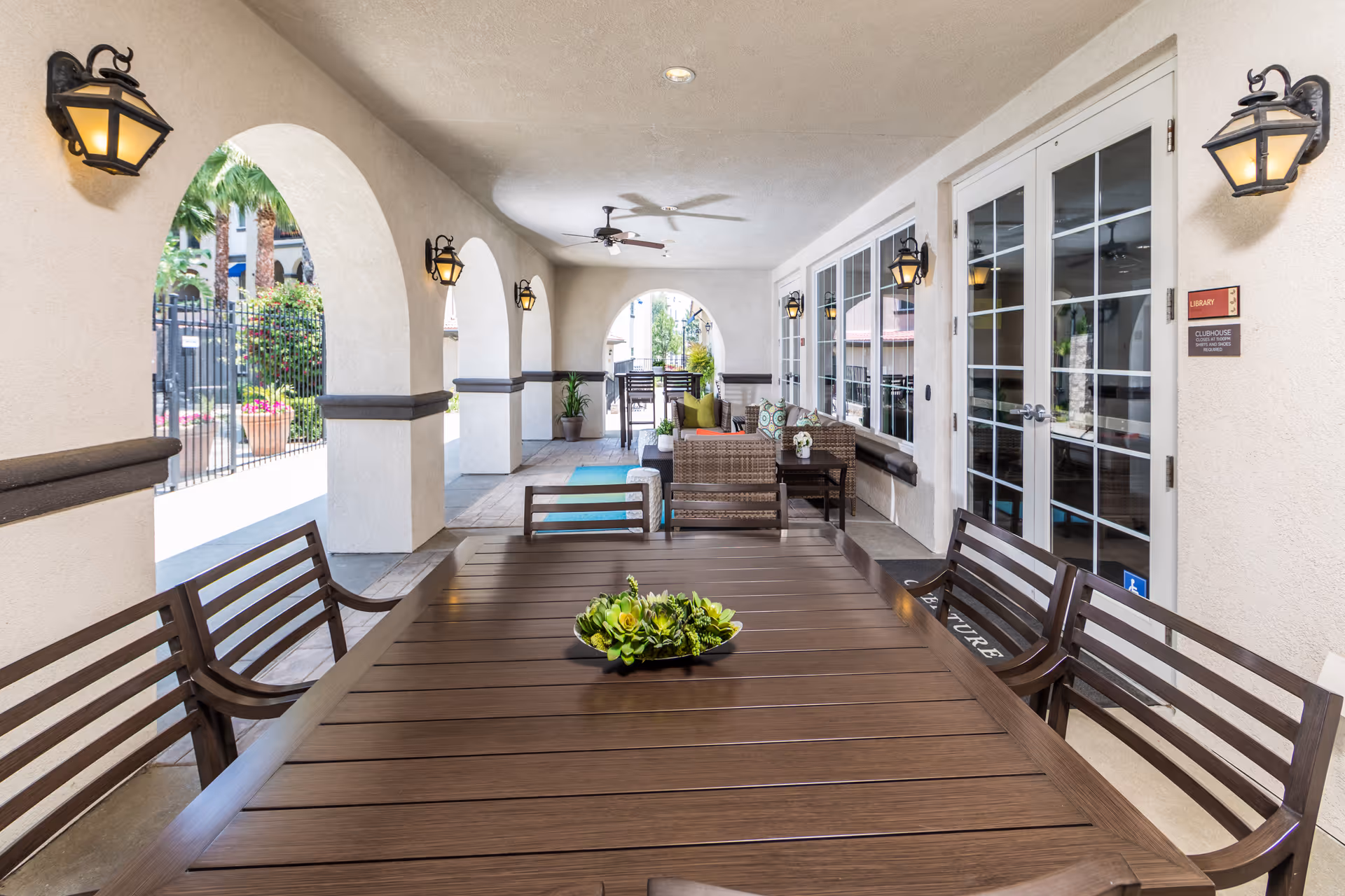 Covered outdoor patio area with a large wooden table and chairs in the foreground, wicker seating with cushions in the background, ceiling fans, wall-mounted lantern lights, and arched openings showing greenery outside.