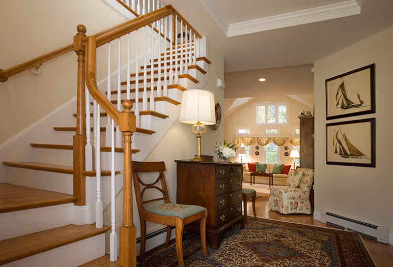 Entryway with a wooden staircase, a small dresser and lamp, chairs, and a view into a sunlit living room.